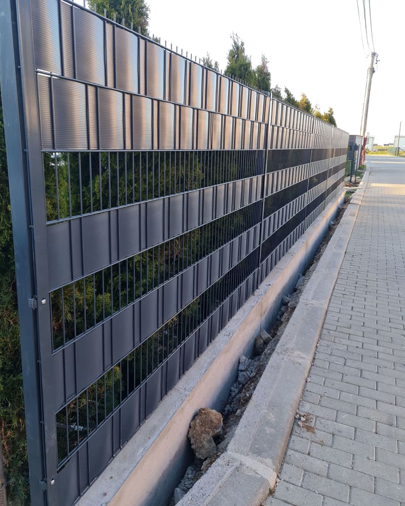 Gardh i gjatë me panele antracit 2D me shirita gri privatësie, përgjatë një trotuari me kalldrëm